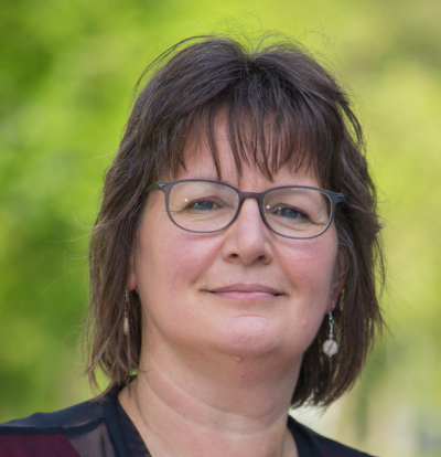 Headshot of Jackie Benschop, a woman with brown hair and glasses in front of a green background.